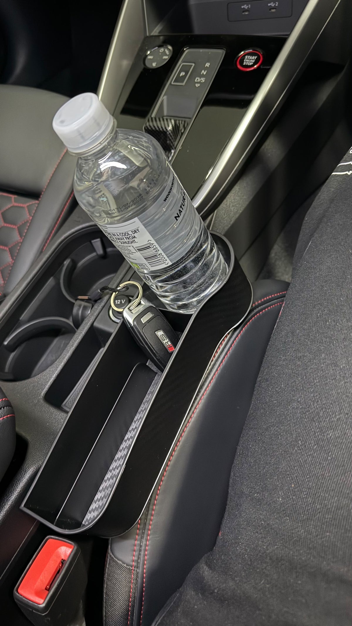 The Car Edit Gap Keeper fitted in the seat gap of a car, holding a water bottle in the cup holder and keys in the tray